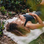 A woman with curly hair meditates on a white towel amidst greenery outdoors, embracing nature.
